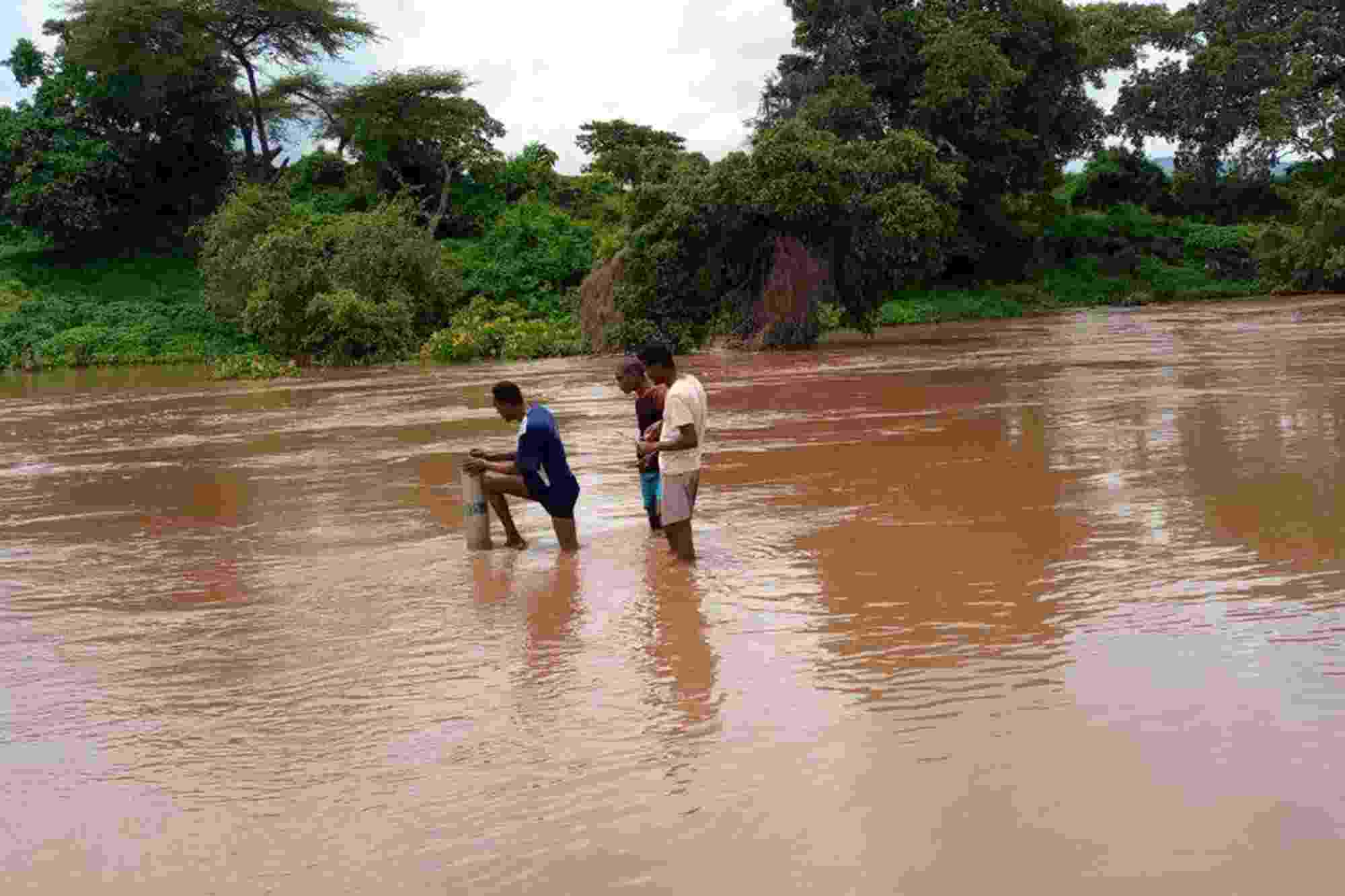 Three men stand knee-deep in brown coloured water as they inspect a water monitoring station, with lush greenery lining the bank behind them